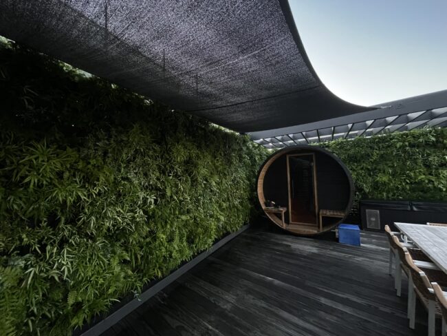 Outdoor seating area with wooden deck, circular pod lounge, and dense green hedge under a black shade sail.