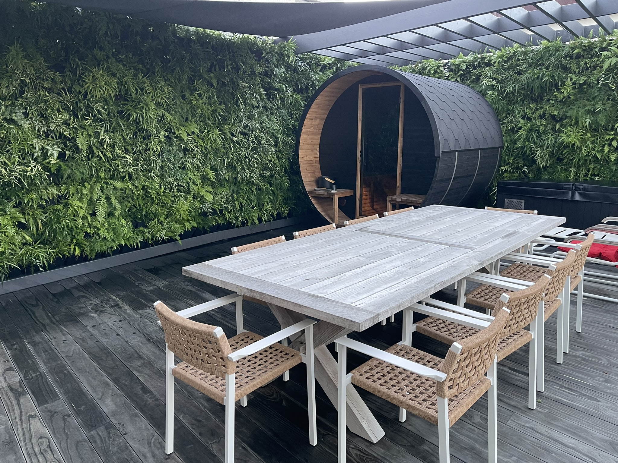 Outdoor dining area with long wooden tables and woven chairs facing a green hedge wall; a round black sauna pod sits in the background.